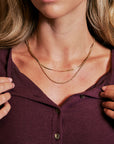 A close-up of a woman wearing a double-layered gold necklace over a dark plum button-up shirt.