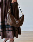 A person wearing a brown tiered maxi dress and clear high-heeled sandals holds a brown snakeskin-patterned crescent shoulder bag with a black strap and gold hardware.