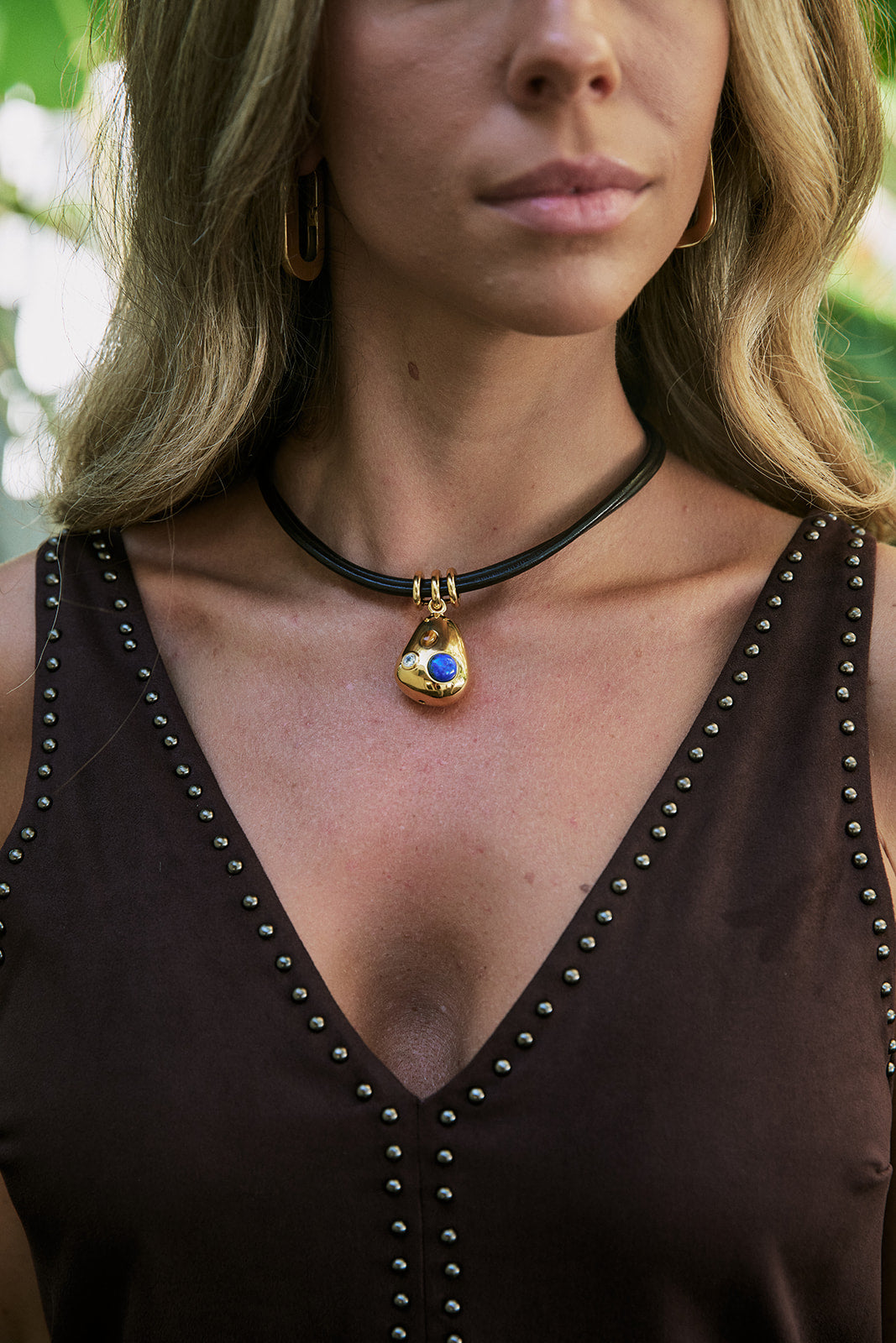 A close-up of a woman wearing a black choker necklace with a gold teardrop pendant featuring a blue evil eye design, paired with a brown studded top.
