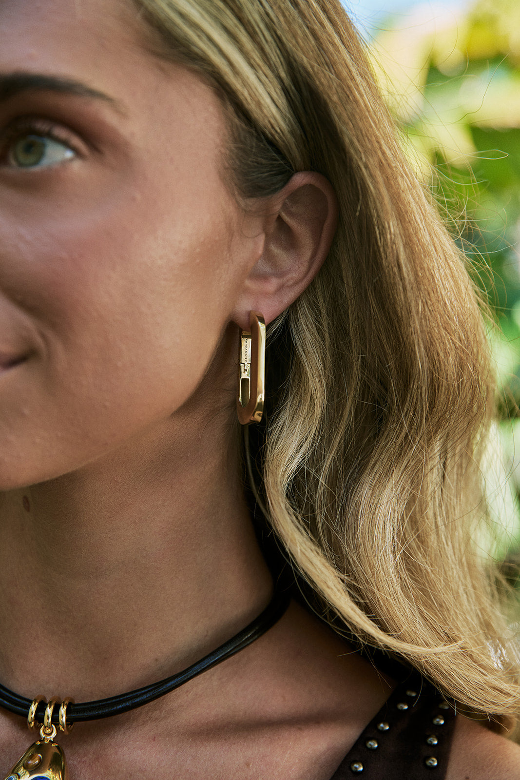 A close-up lifestyle shot of a woman wearing a gold geometric hoop earring, with her blonde hair and ear clearly visible against a soft, blurred green background.