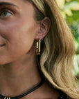 A close-up lifestyle shot of a woman wearing a gold geometric hoop earring, with her blonde hair and ear clearly visible against a soft, blurred green background.