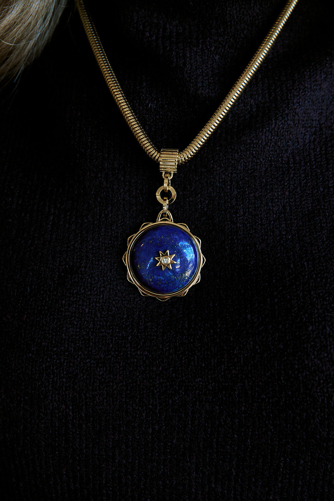 A gold-toned snake chain necklace featuring a circular pendant with a deep blue lapis lazuli-style center and a small gold star motif, displayed on a model wearing a black top