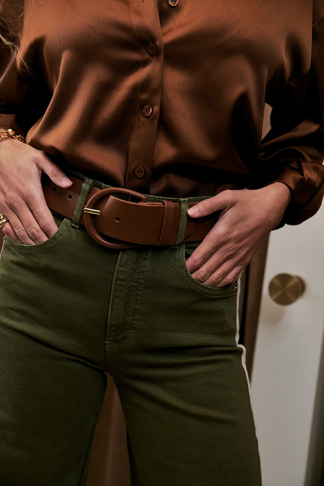 A close-up of a person wearing a brown satin button-up shirt, an olive green pair of pants, and a wide brown leather belt with a large oval buckle.