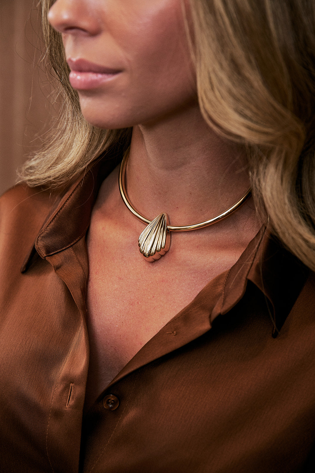 A close-up lifestyle shot of a woman wearing a gold-toned metal necklace with a sculptural, shell-like pendant, paired with a brown satin button-down shirt.