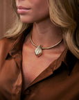 A close-up lifestyle shot of a woman wearing a gold-toned metal necklace with a sculptural, shell-like pendant, paired with a brown satin button-down shirt.