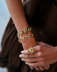 A close-up of a person's wrist and hand wearing multiple gold-tone jewelry pieces, including a spiral ring, a chain bracelet, and a bangle, against a dark brown dress.