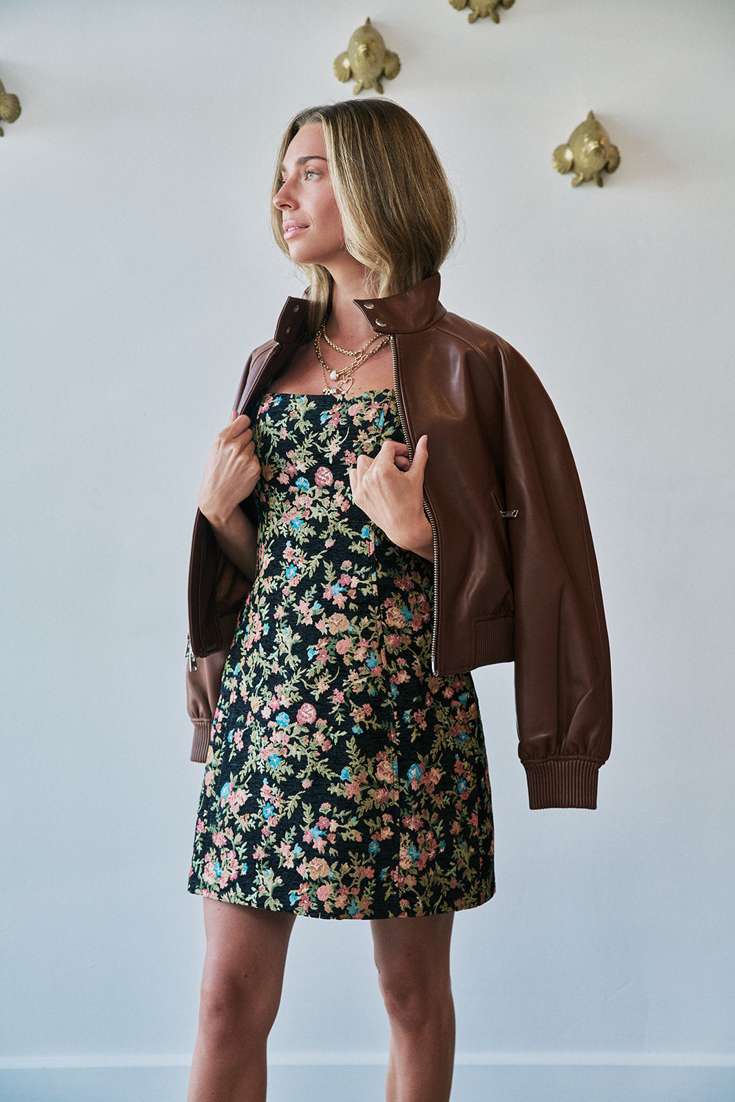 A woman wearing a brown faux-leather bomber jacket over a black floral strapless dress, standing against a white wall with gold wall sconces.