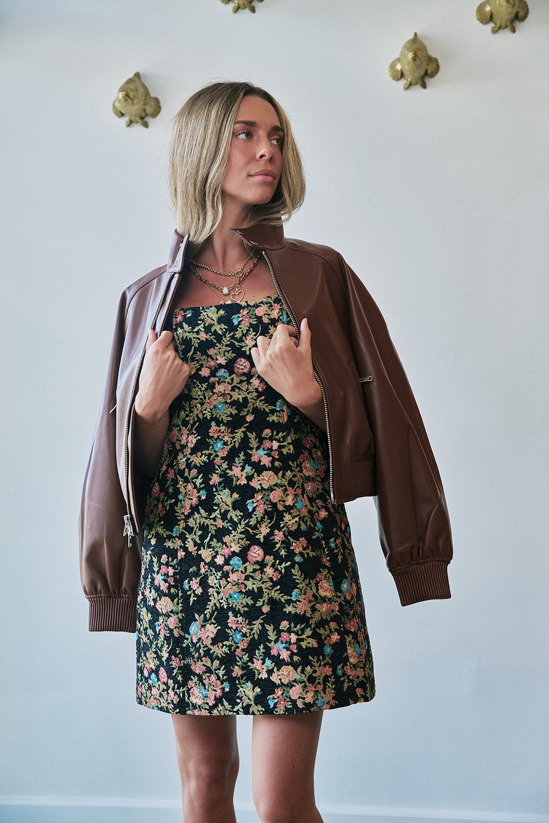 A woman wearing a brown faux-leather bomber jacket over a black floral strapless dress, standing against a light gray wall with gold decorative wall hangings.