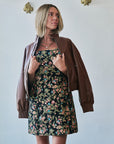 A woman wearing a brown faux-leather bomber jacket over a black floral strapless dress, standing against a light gray wall with gold decorative wall hangings.