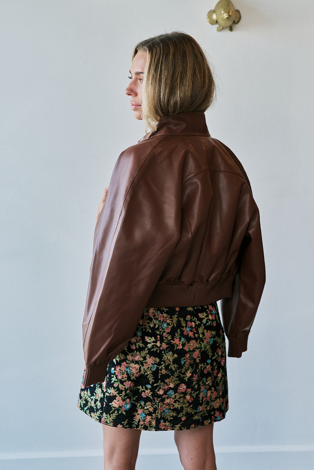 A woman wearing a brown leather cropped jacket and a black floral mini skirt, shown from a side-back angle against a plain light gray wall.