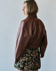 A woman wearing a brown leather cropped jacket and a black floral mini skirt, shown from a side-back angle against a plain light gray wall.
