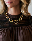 A close-up of a woman wearing a chunky gold chain necklace with large interlocking links, paired with a dark brown top.