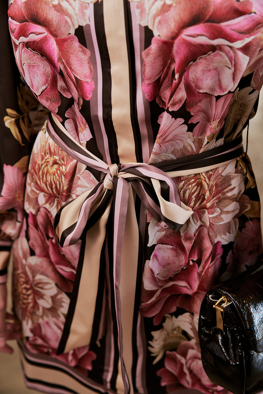 A close-up of a floral dress or garment featuring large pink and mauve peony-like flowers over a black base with vertical pink and beige stripes, tied at the waist with a matching satin ribbon. A small black handbag is partially visible at the lower right.