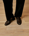 A close-up of a person's feet wearing dark brown crocodile-embossed loafers with black trousers, standing on a light-colored wooden floor.
