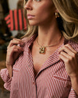 A close-up lifestyle shot of a woman wearing a gold necklace with a sculptural, abstract animal-head pendant. She is wearing a red and white vertically striped button-down shirt, with a softly blurred boutique background.