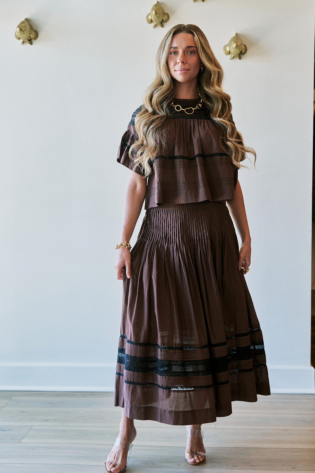 A woman modeling a brown two-piece dress with a tiered short-sleeve top and a pleated maxi skirt, featuring black horizontal stripes near the hem and shoulders, set against a white wall with gold wall decor.