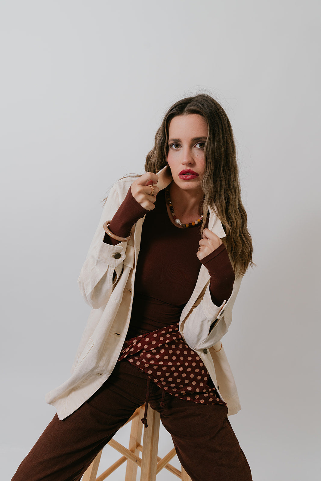 Woman in ivory jacket, brown bodysuit, polka dot scarf and brown pants against a plain background.