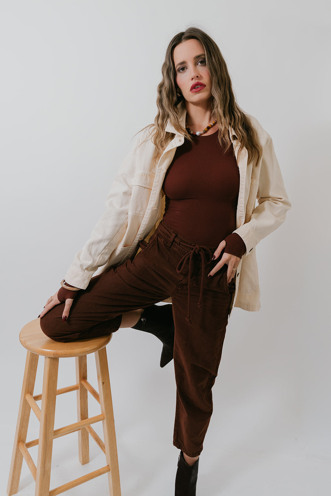 Woman in brown outfit sitting on a wooden stool against a white background