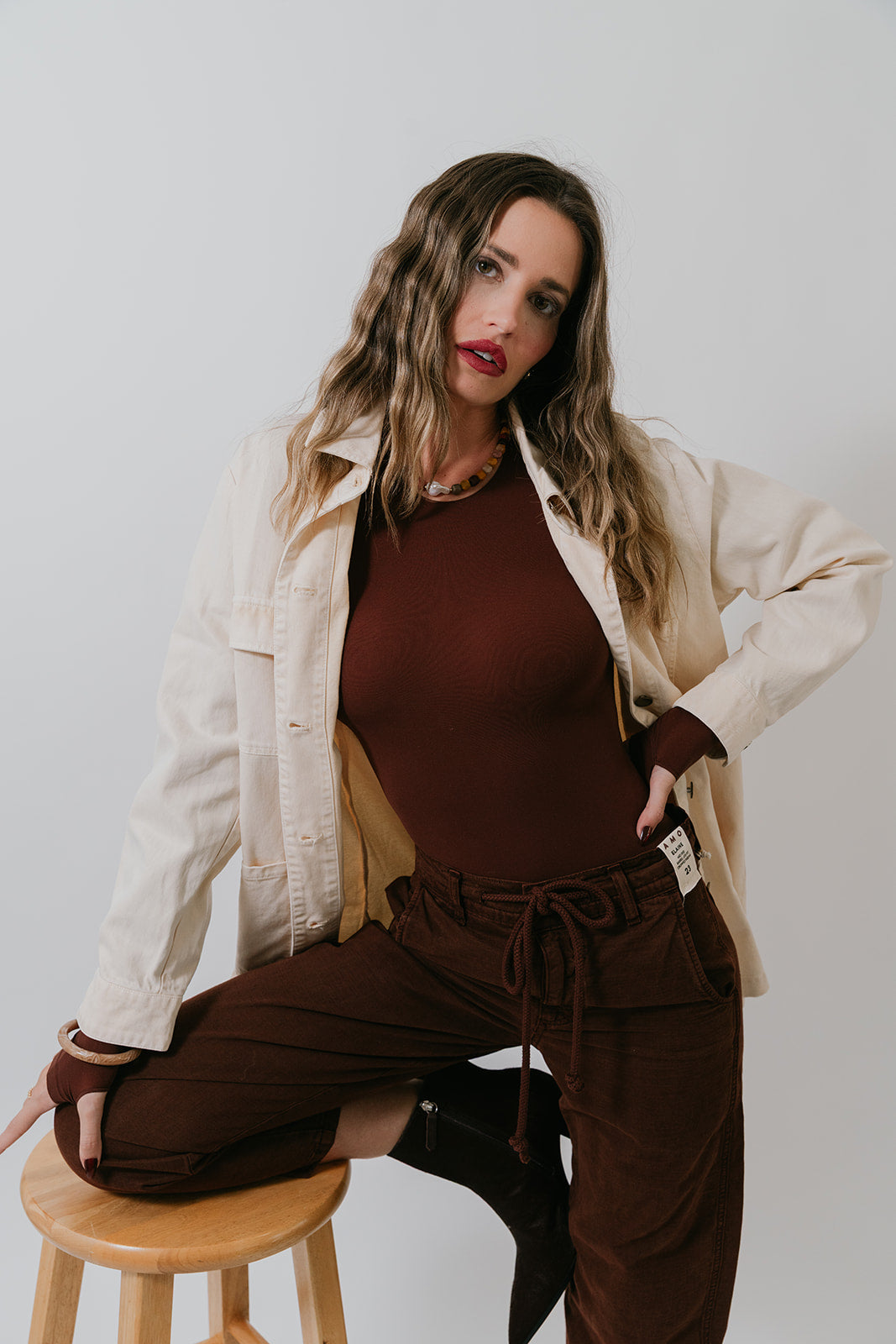Woman in a beige jacket, Ballet Crewneck Bodysuit and brown pants sitting on a stool against a plain background