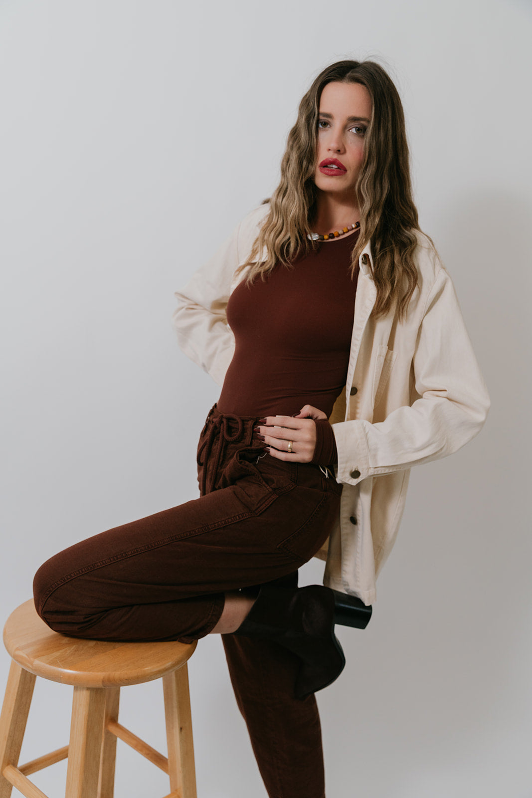 Woman sitting on a wooden stool wearing a brown outfit with a Charlie Chore Coat against a plain background