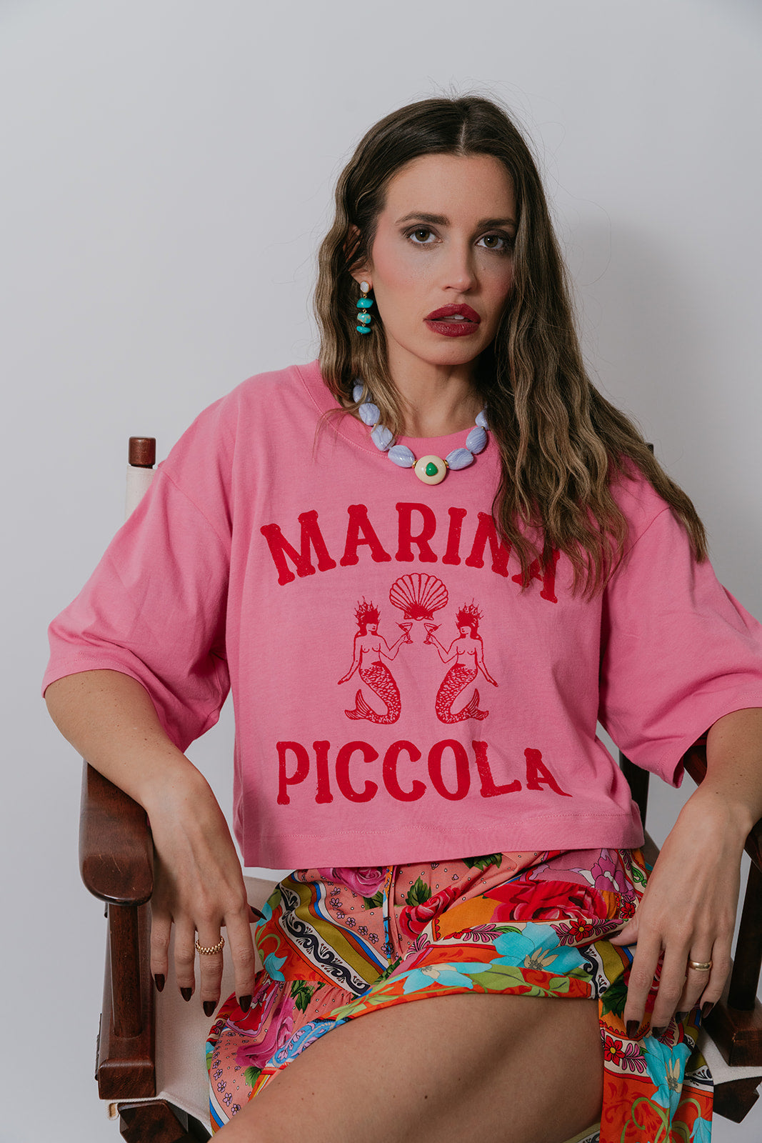 Woman wearing a pink 'Marina Piccola' shirt sitting on a chair.