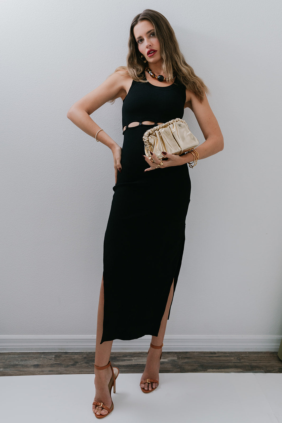 Woman in a black dress holding a gold clutch against a white wall