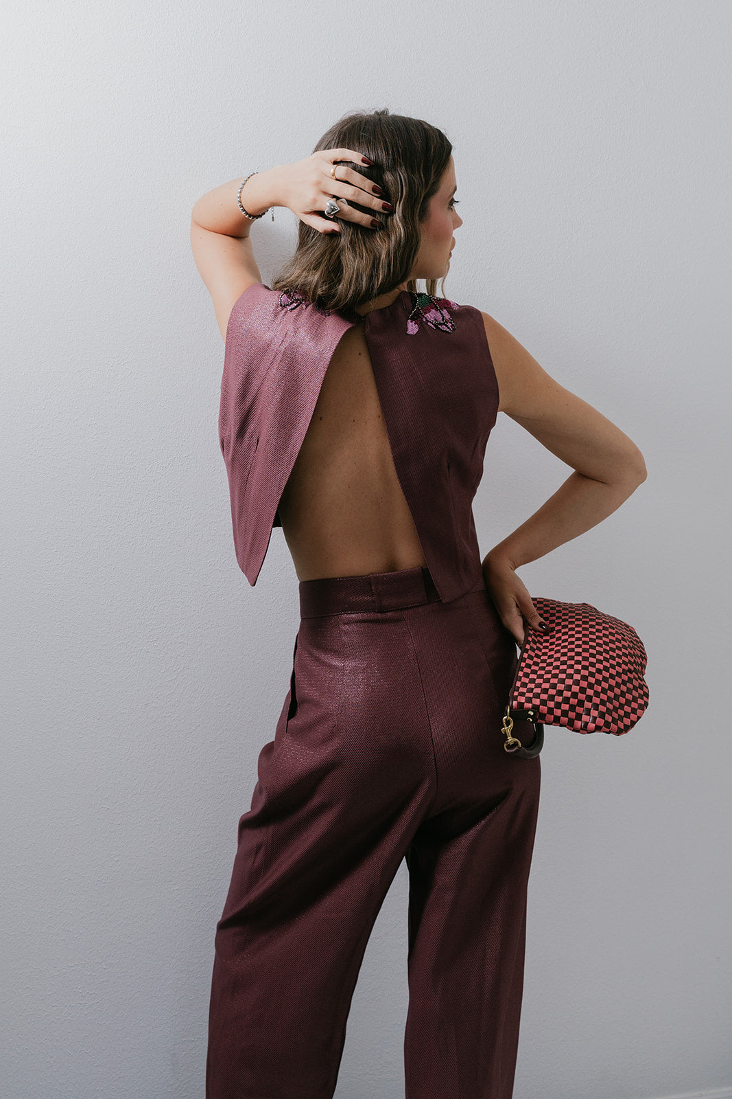 Woman in a two-piece outfit with a maroon top and maroon pants, holding a checkered clutch against a plain background. back view