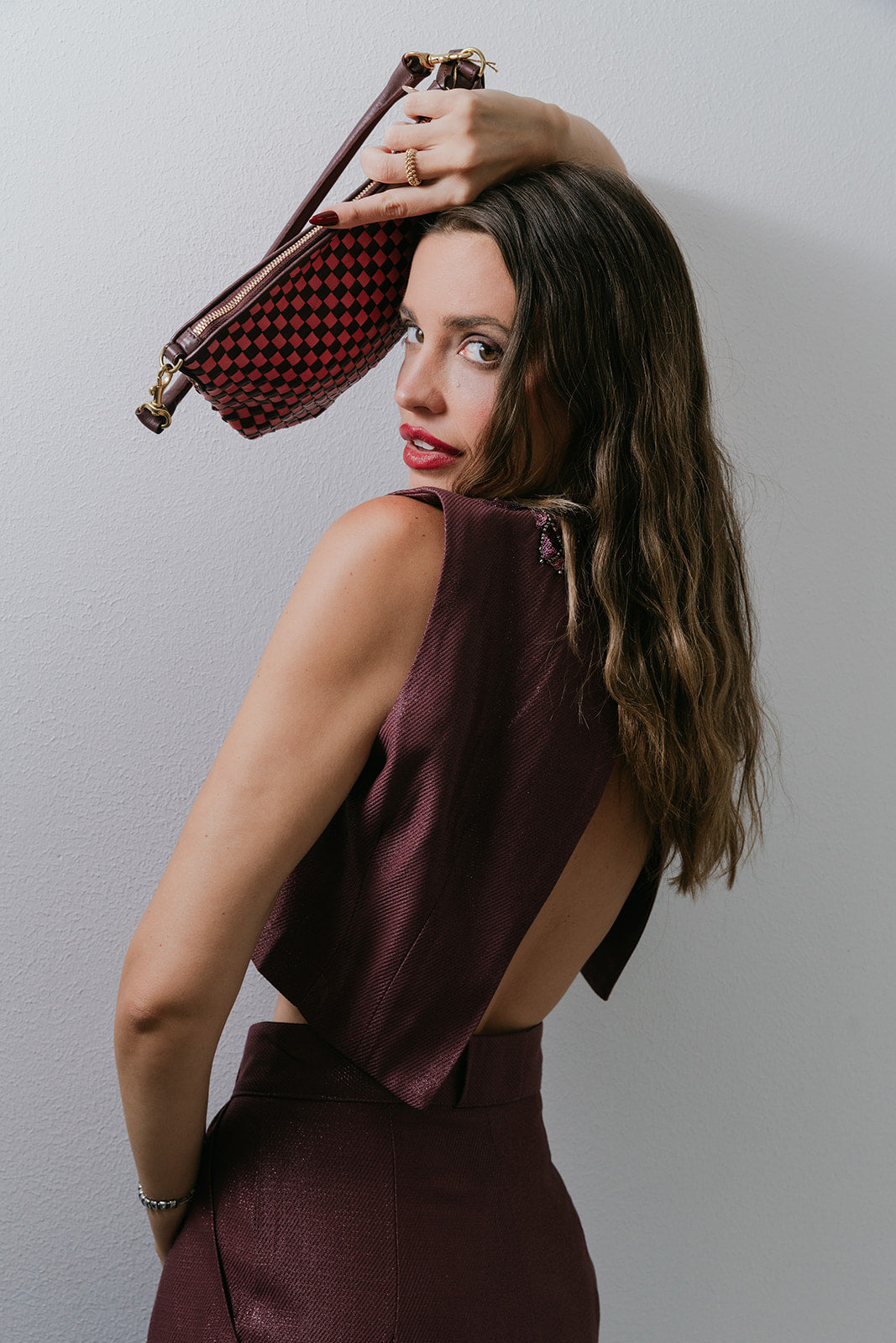 Woman in a burgundy set holding a checkered handbag against a plain background, back view