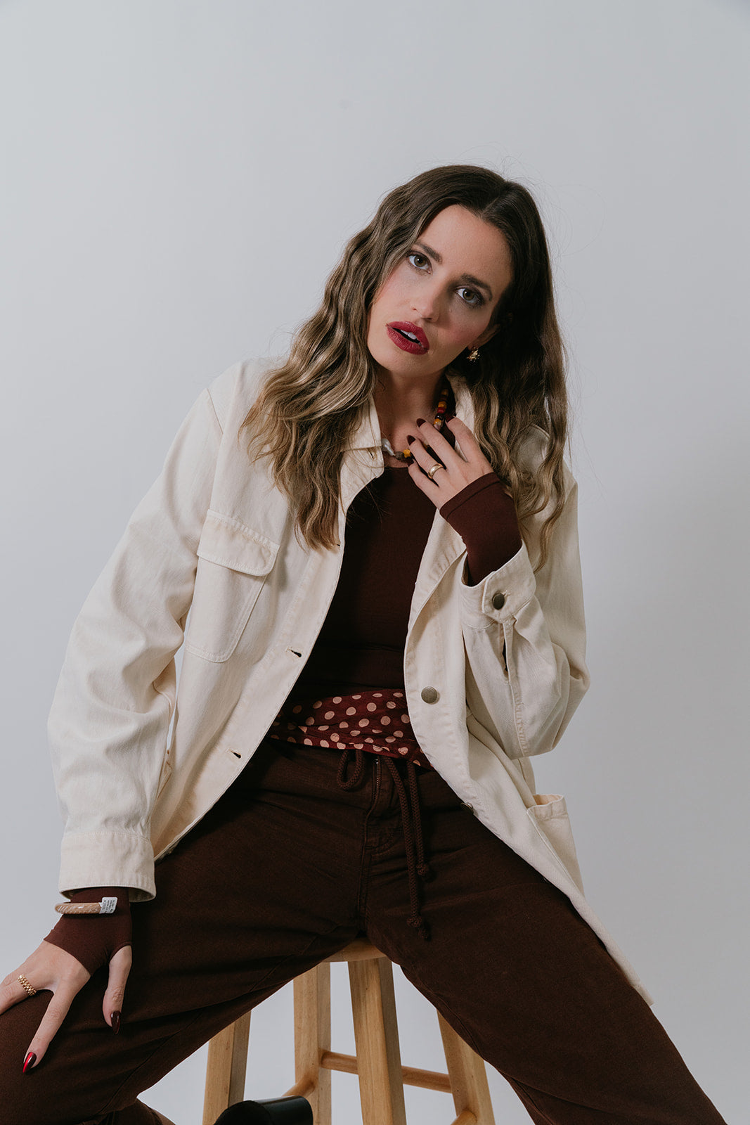 Woman sitting on a stool wearing a Charlie Chore Coat and brown pants against a plain background