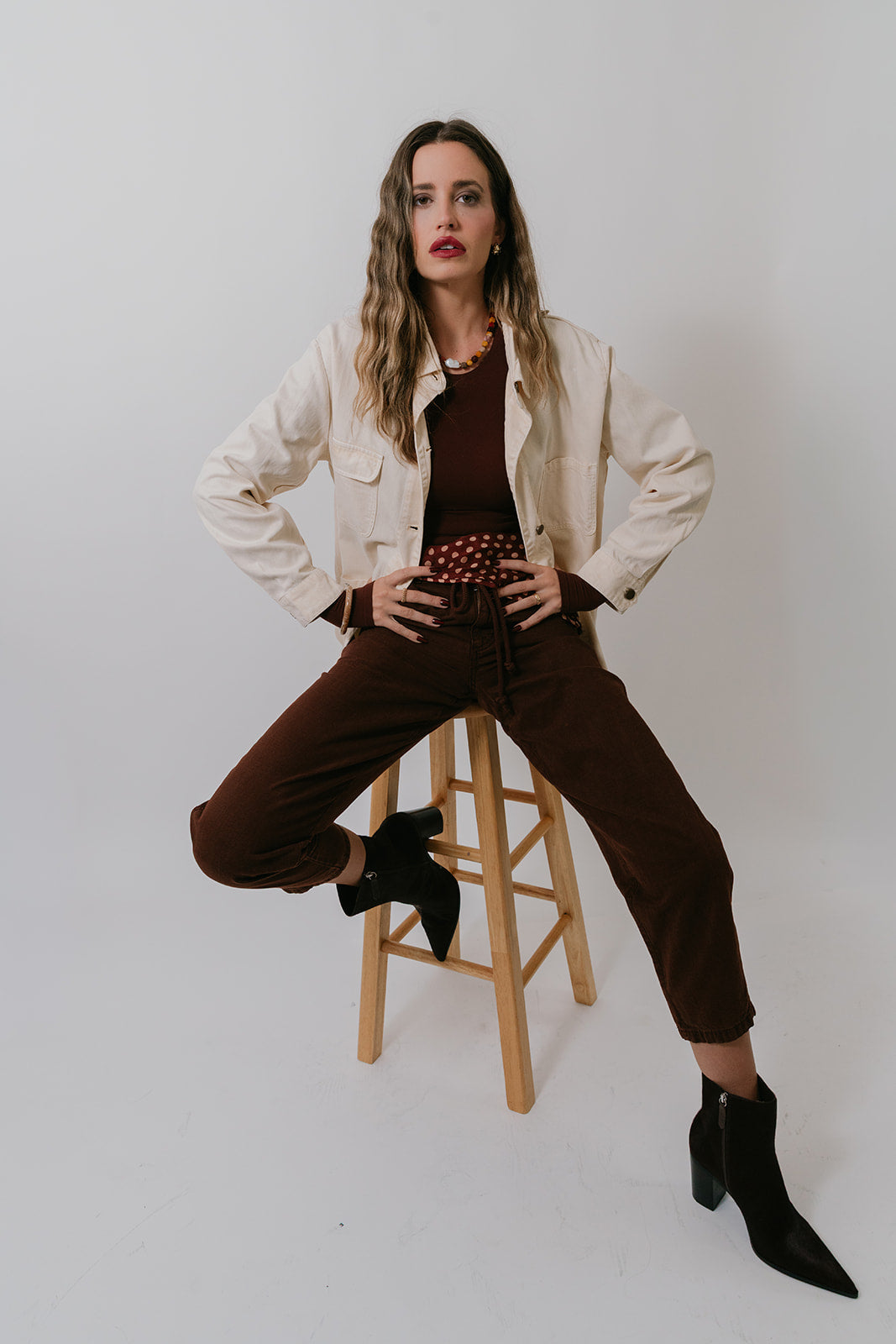 Woman sitting on a stool wearing a beige jacket, brown pants, and black shoes against a white background