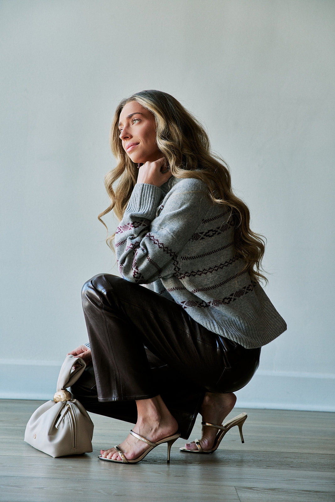 A woman crouches in a studio setting wearing a gray knit sweater with a dark geometric pattern, dark brown pants, and metallic heeled sandals, while holding a small cream-colored handbag.
