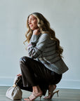 A woman crouches in a studio setting wearing a gray knit sweater with a dark geometric pattern, dark brown pants, and metallic heeled sandals, while holding a small cream-colored handbag.