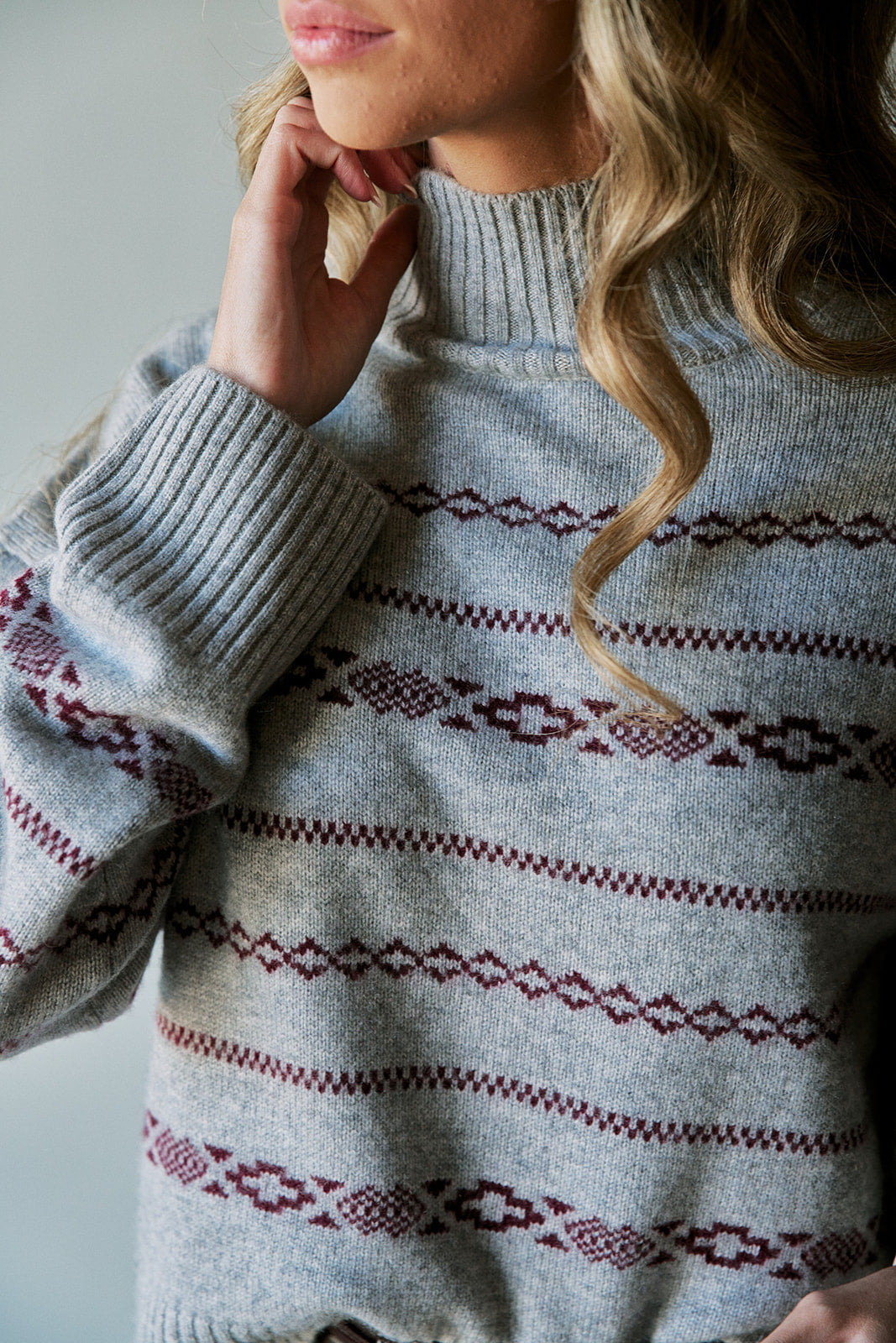 A close-up of a woman wearing a light gray knit sweater with burgundy geometric Fair Isle patterns across the chest and sleeves.