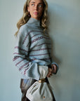 A woman wearing a light blue-gray knit turtleneck sweater with burgundy geometric patterns, paired with dark brown leather pants and holding a cream-colored drawstring handbag, standing against a plain light blue-gray wall.