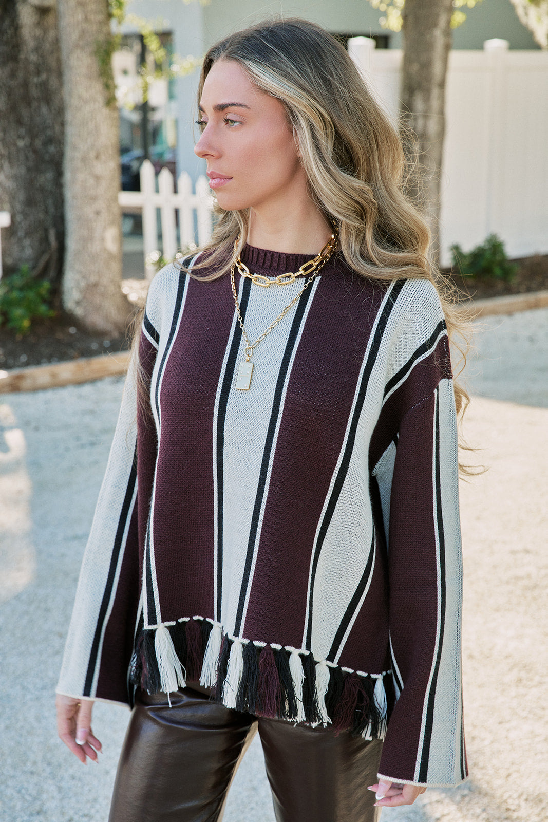 A woman wearing a knit sweater with vertical stripes in burgundy, cream, and black, featuring a fringed hem and long sleeves, standing outdoors.