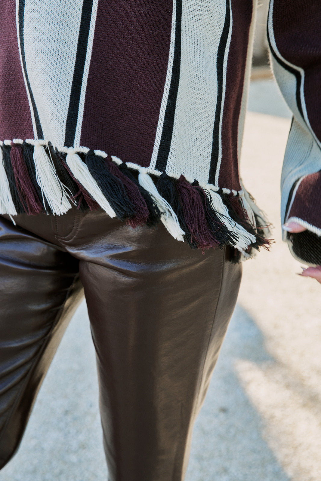 Close-up of a person wearing a burgundy, cream, and black striped knit top with fringe trim, paired with dark brown faux-leather pants, outdoors on a light pavement background.