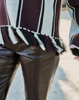 Close-up of a person wearing a burgundy, cream, and black striped knit top with fringe trim, paired with dark brown faux-leather pants, outdoors on a light pavement background.