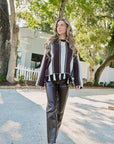 A woman walking outdoors on a gravel path wearing a burgundy, white, and black striped long-sleeve top with a ruffled hem, paired with faux-leather pants and white heeled sandals.