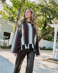 A woman wearing a burgundy and cream striped tassel-trimmed top with faux-leather pants, standing outdoors in front of trees and a white fence.
