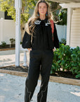 A woman stands outdoors in front of a house porch wearing a black long-sleeve top with white swirl embroidery and matching black pants with white line details, paired with brown boots.