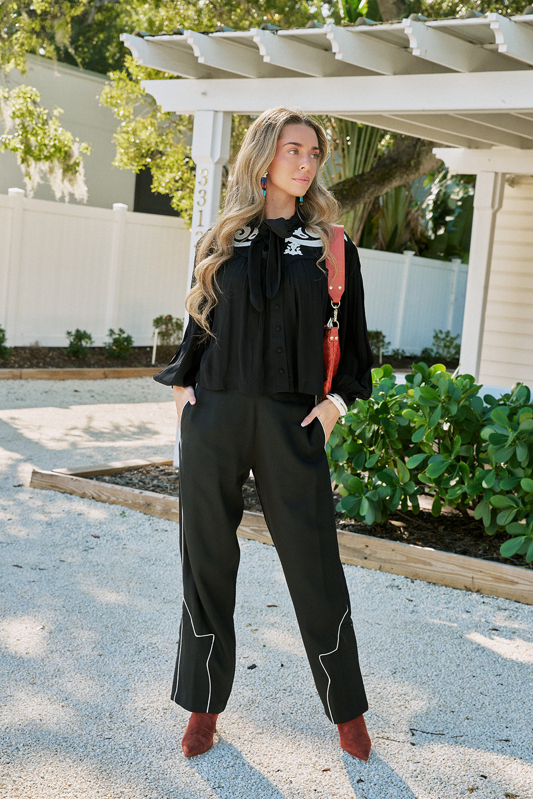 A woman stands outdoors in a garden courtyard wearing a black long-sleeve top and matching black wide-leg pants with white contrast piping, paired with brown boots and a red shoulder bag.