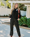A woman stands outdoors in a garden courtyard wearing a black long-sleeve top and matching black wide-leg pants with white contrast piping, paired with brown boots and a red shoulder bag.
