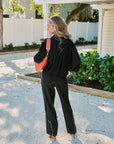 A woman stands outdoors on a gravel driveway wearing a black long-sleeve top and black wide-leg pants, with a bright coral-orange shoulder bag and matching orange shoes. A white house, greenery, and pergola are visible in the background.
