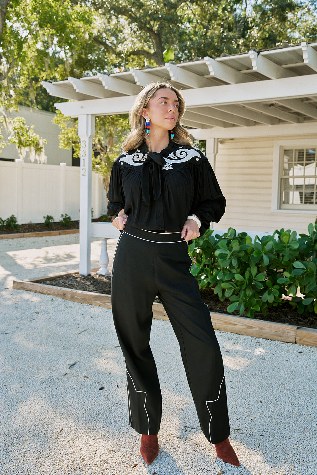 A woman modeling a black long-sleeve blouse and matching wide-leg trousers with white western-style embroidery and piping, standing outdoors on a gravel driveway in front of a white house and greenery.