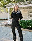 A woman modeling a black long-sleeve blouse and matching wide-leg trousers with white western-style embroidery and piping, standing outdoors on a gravel driveway in front of a white house and greenery.