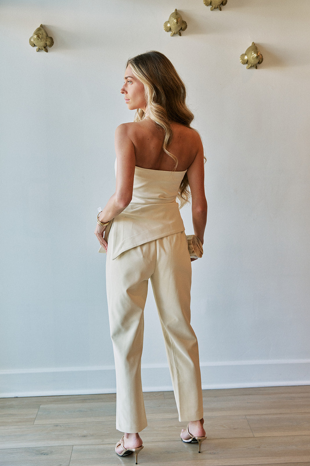 A woman stands with her back to the camera wearing a light beige strapless tube top and matching wide-leg trousers, paired with metallic heels, in a minimalist room with a white wall and gold wall art.