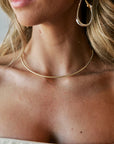 A close-up lifestyle shot of a woman wearing a delicate gold snake chain necklace and a large gold hoop earring.