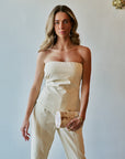 A woman modeling a strapless cream-colored top and matching pant with a structured bodice and straight legs, holding a small patterned clutch against a light wall.