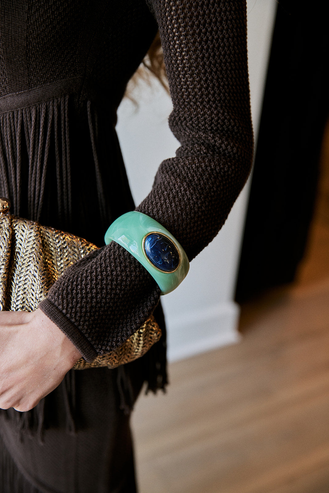 A close-up of a person wearing a wide mint green cuff bracelet with a dark blue oval stone, paired with a dark brown textured knit top and holding a gold patterned clutch.