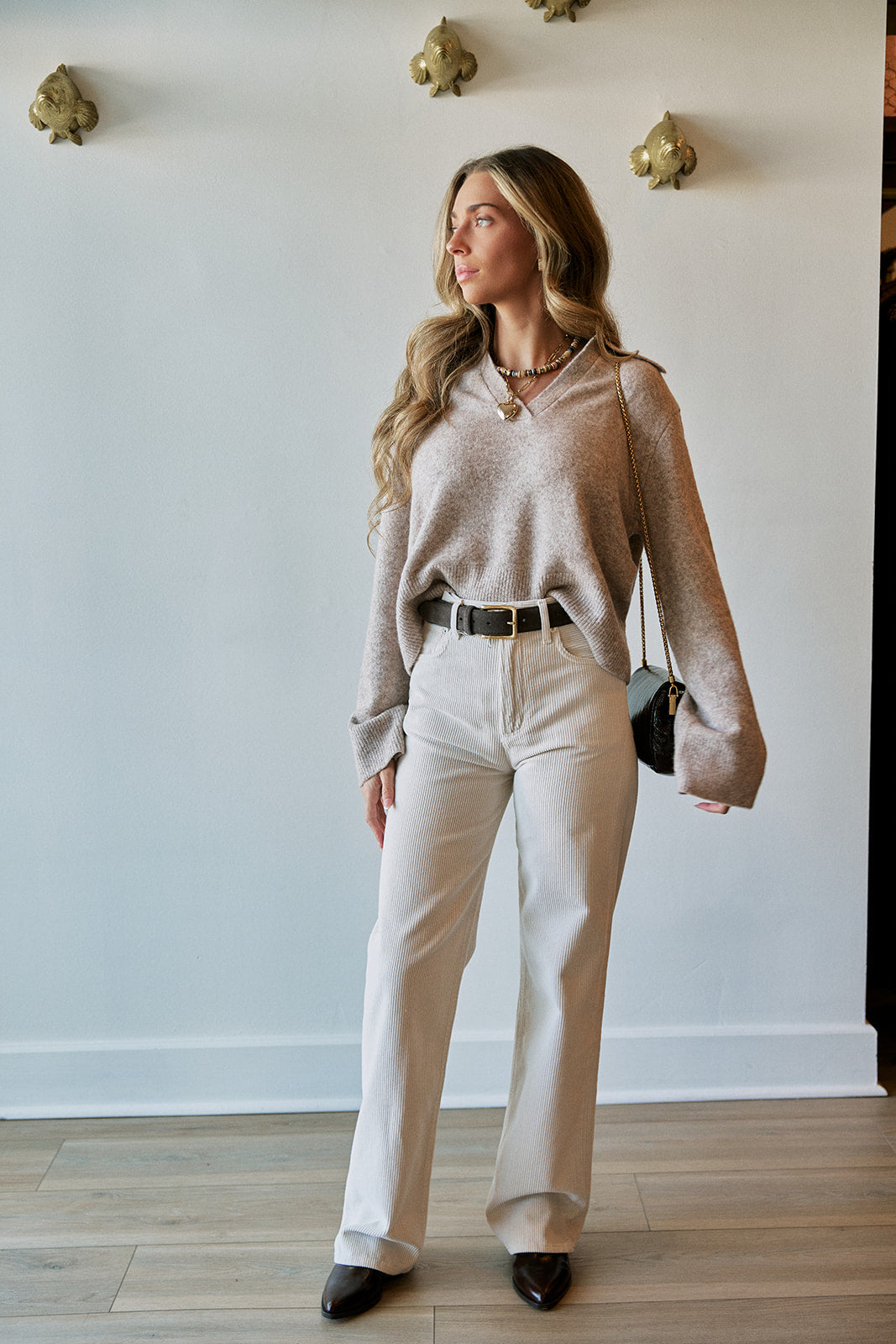 A woman stands in a room wearing a shimmering champagne long-sleeve knit sweater, off-white wide-leg trousers, a black belt, and black pointed shoes. She is holding a small black handbag and wearing layered gold necklaces.